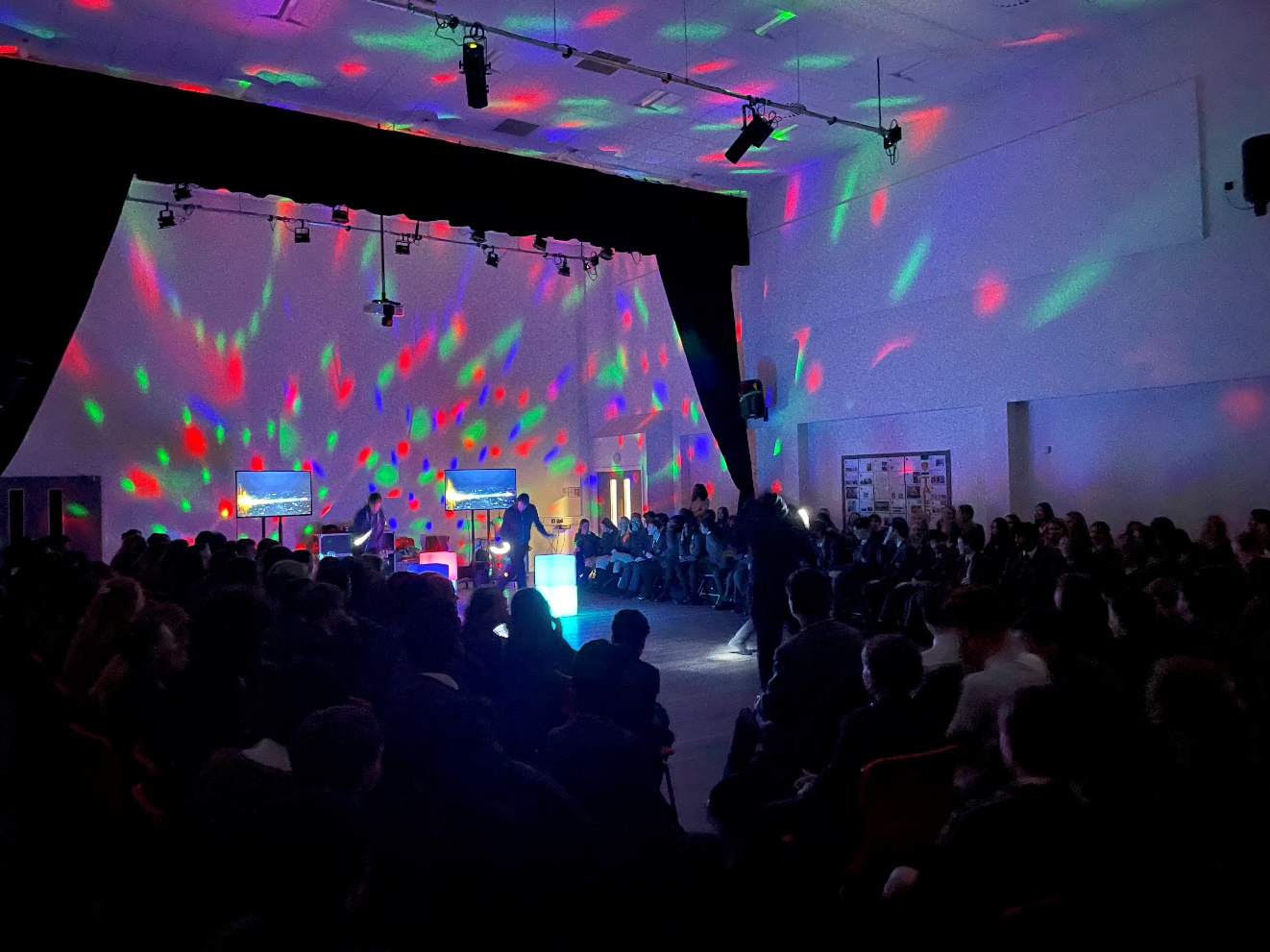 A crowd of people in a room with a stage and a stage with lights

Description automatically generated